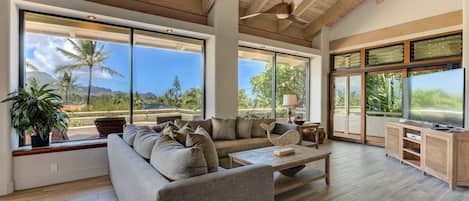A spacious living room with a large sofa, a coffee table, and a television. There are large windows offering a view of palm trees and mountains.