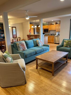 A living room with a sofa, armchairs, and a coffee table. There is a kitchen in the background with wooden cabinets and appliances.