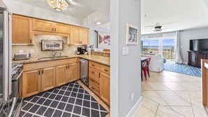 Cocina moderna con armarios de madera, un refrigerador de acero inoxidable y una encimera de granito. Hay un área de comedor con un sofá blanco y una televisión. La habitación tiene vista a la playa.