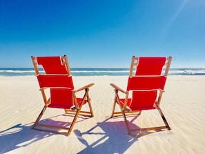 Zwei rote Liegestühle an einem Sandstrand, im Hintergrund das Meer.