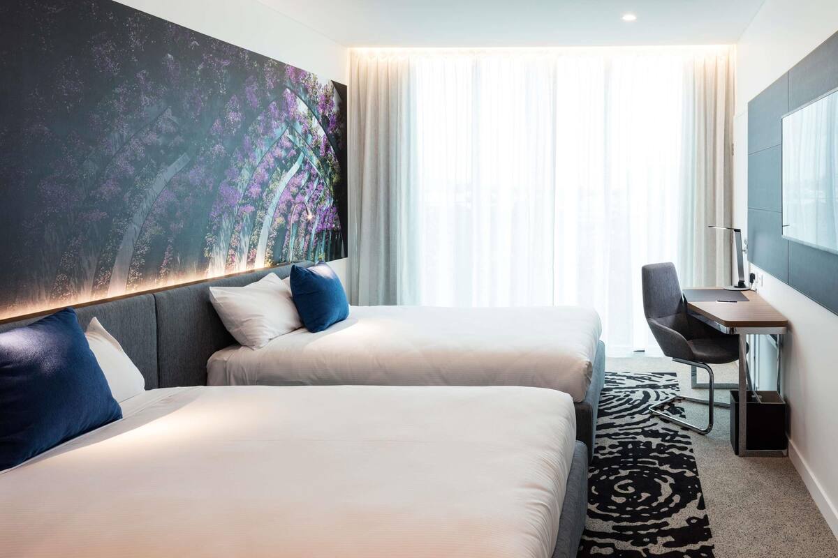 Habitación de hotel con dos camas, un escritorio, una silla, un cuadro abstracto grande y vistas de la ciudad a través de cortinas traslúcidas.