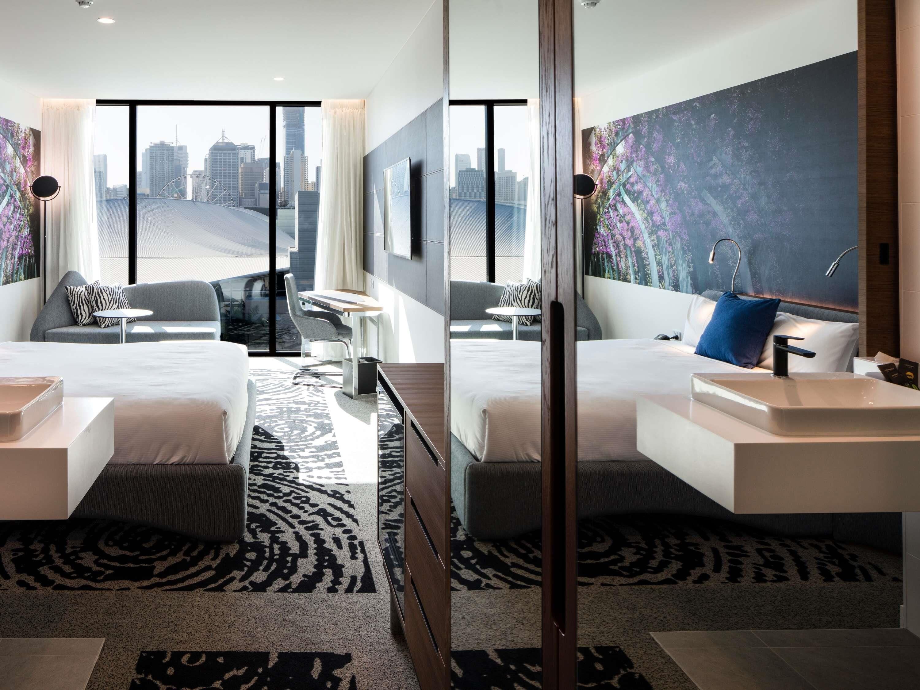 A modern hotel room with a city view, a large bed, two seating areas, and a bathroom with a sink and mirror.