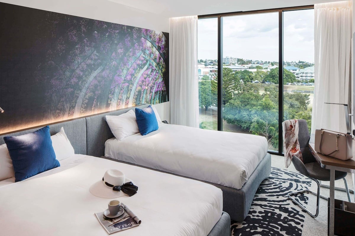 Habitación de hotel con dos camas, un ventanal con vistas a la ciudad, un cuadro en la pared y una mesita con una silla.