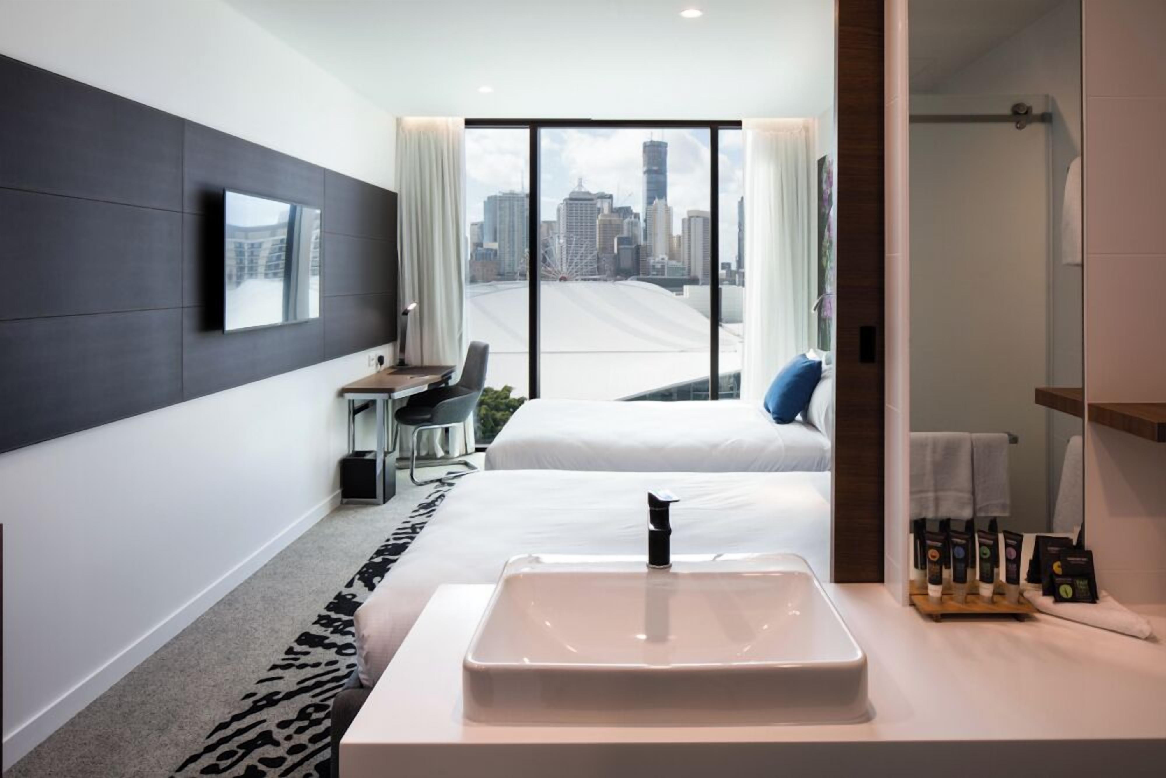 Una habitación de hotel moderna con una cama grande, un lavamanos blanco, un escritorio con una silla y vistas a la ciudad a través de amplios ventanales.