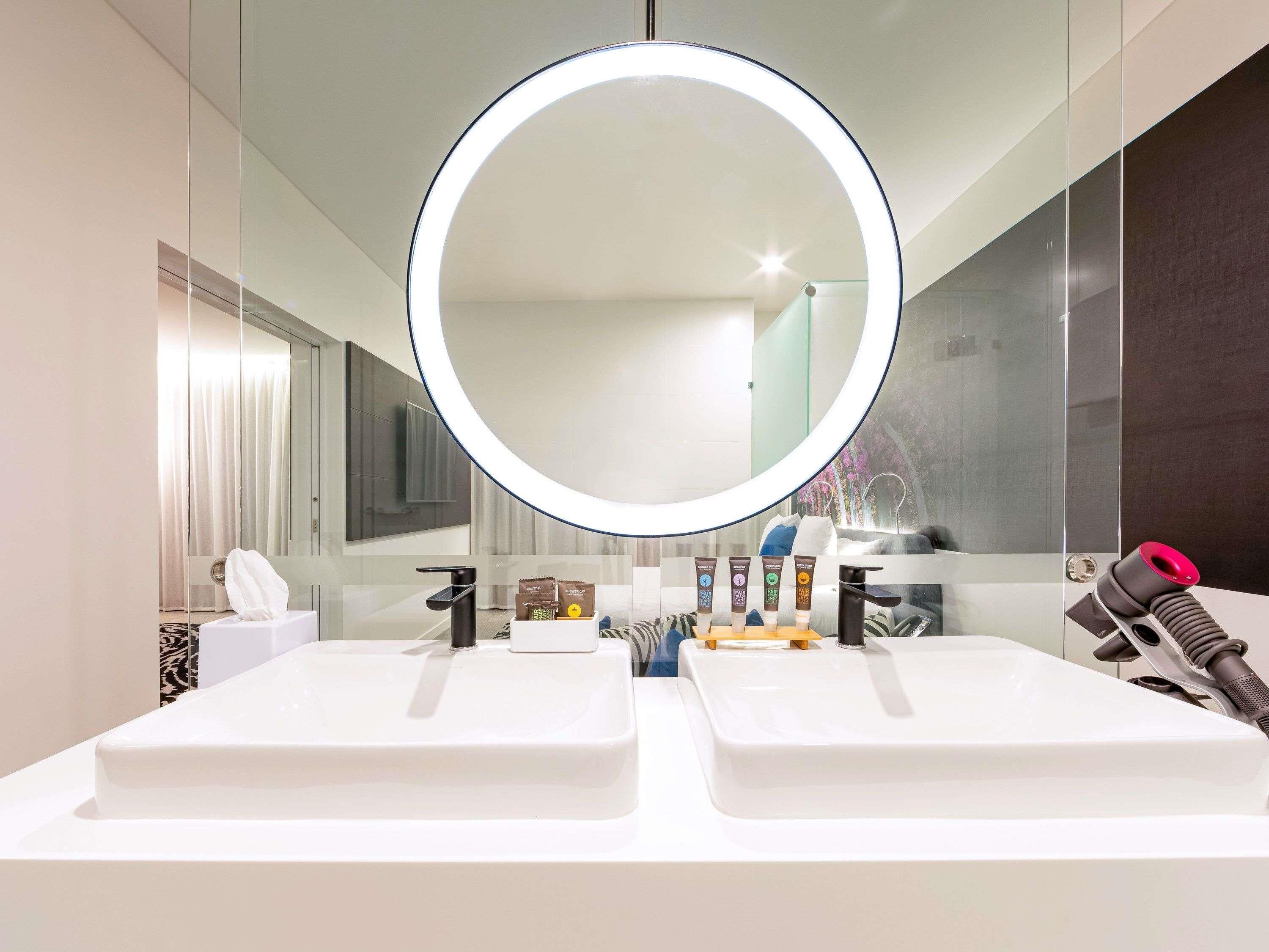 Une salle de bain moderne avec deux lavabos, un grand miroir avec éclairage intégré et un sèche-cheveux placé à droite.