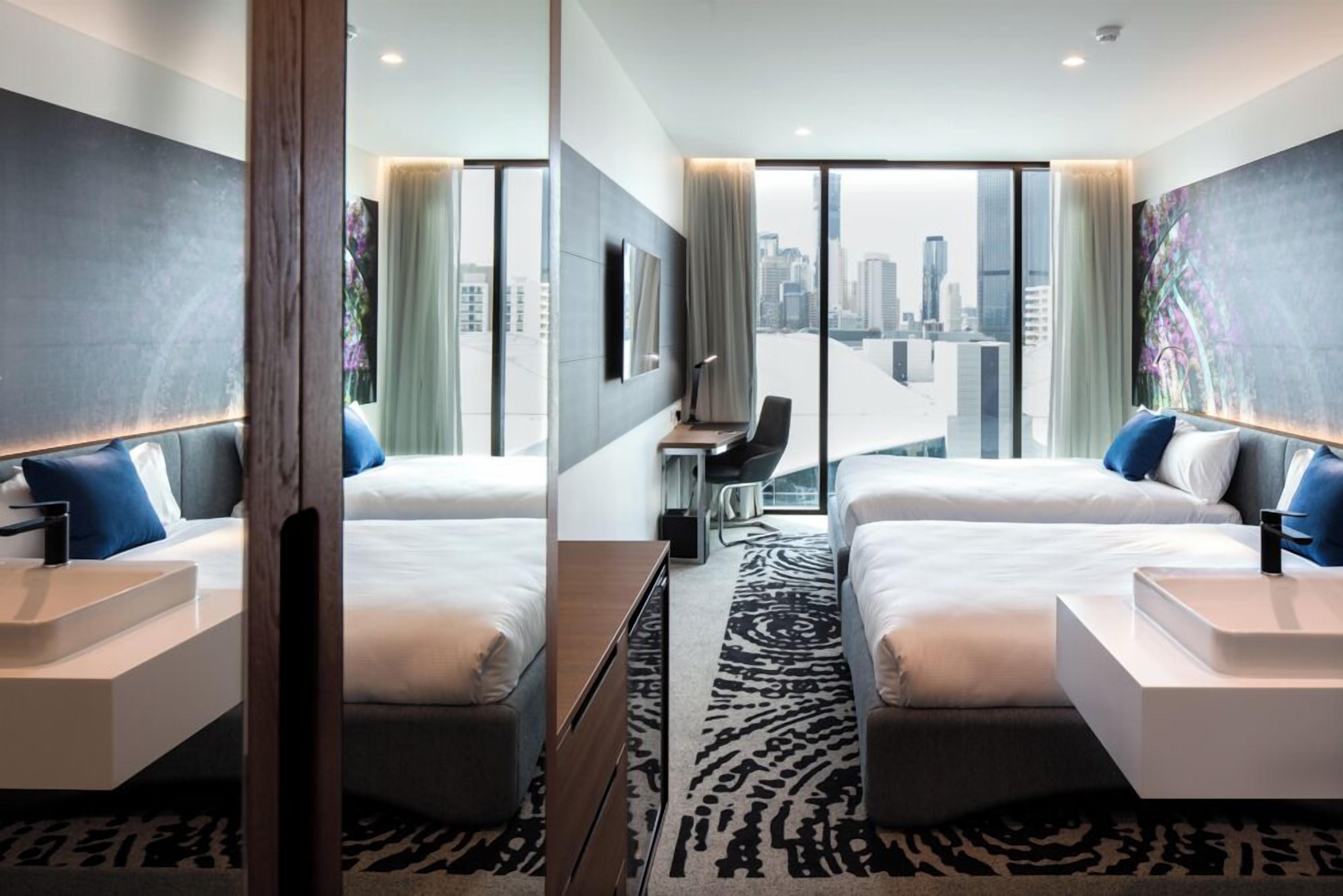 Une chambre d’hôtel moderne avec deux lits, un bureau et une grande fenêtre donnant sur la ville.