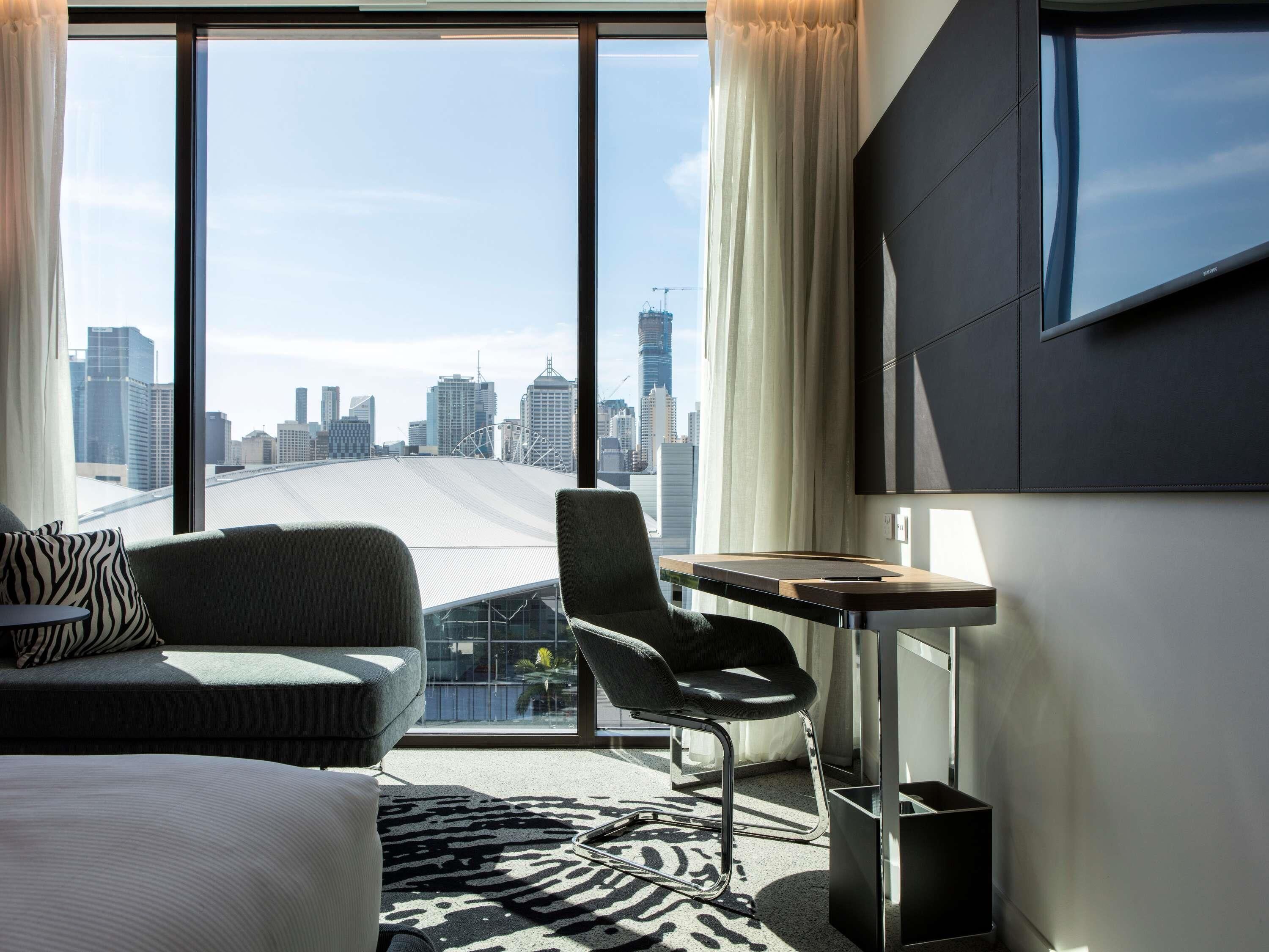 A hotel room with a large window, a sofa, a chair, a small table, and a TV mounted on the wall.