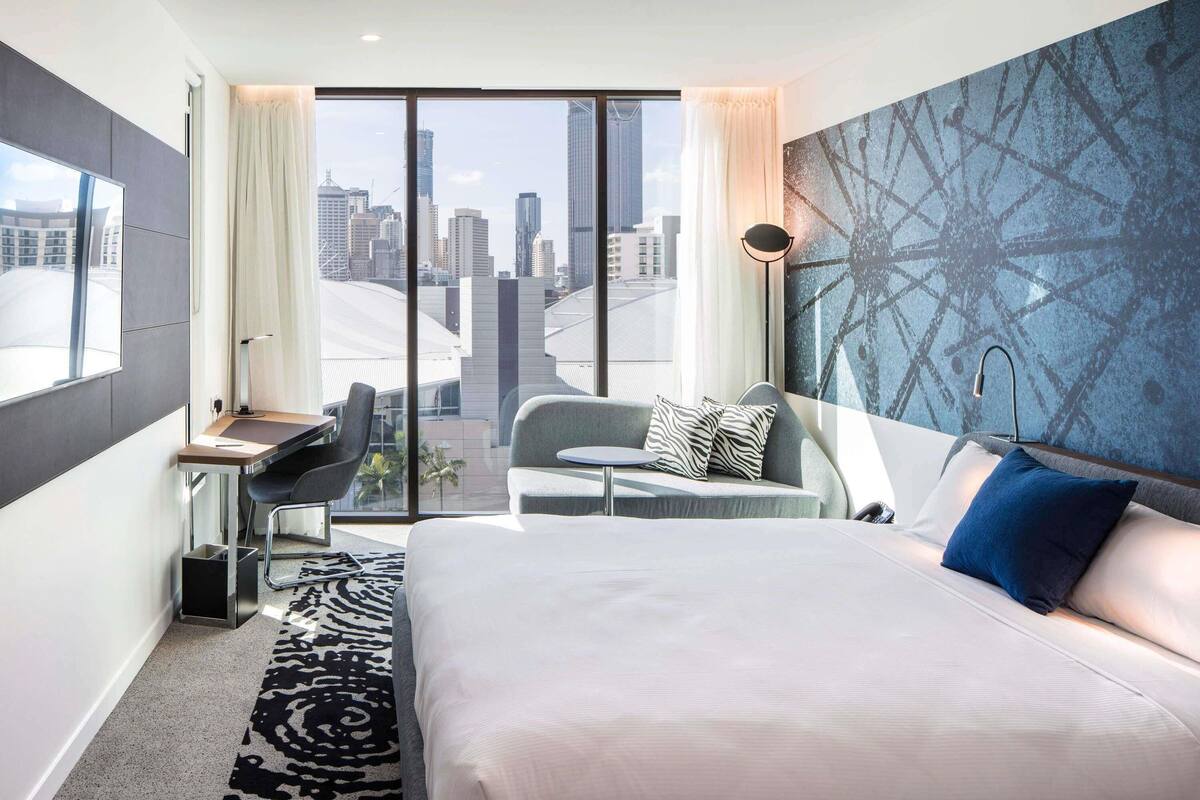 Habitación de hotel moderna con una cama grande, un escritorio, una silla y vistas a la ciudad a través de ventanales de pared a techo.