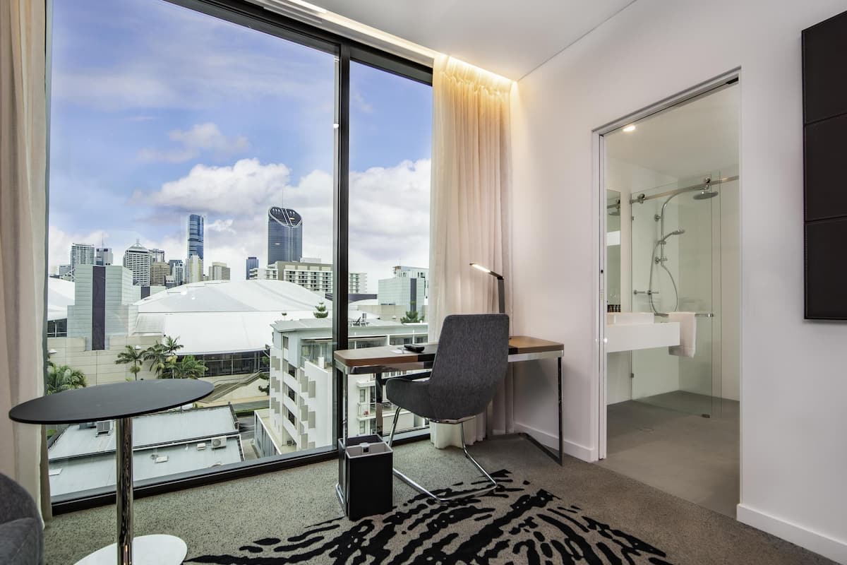 A modern hotel room with a city view, a desk, a chair, a glass shower, and a large window.