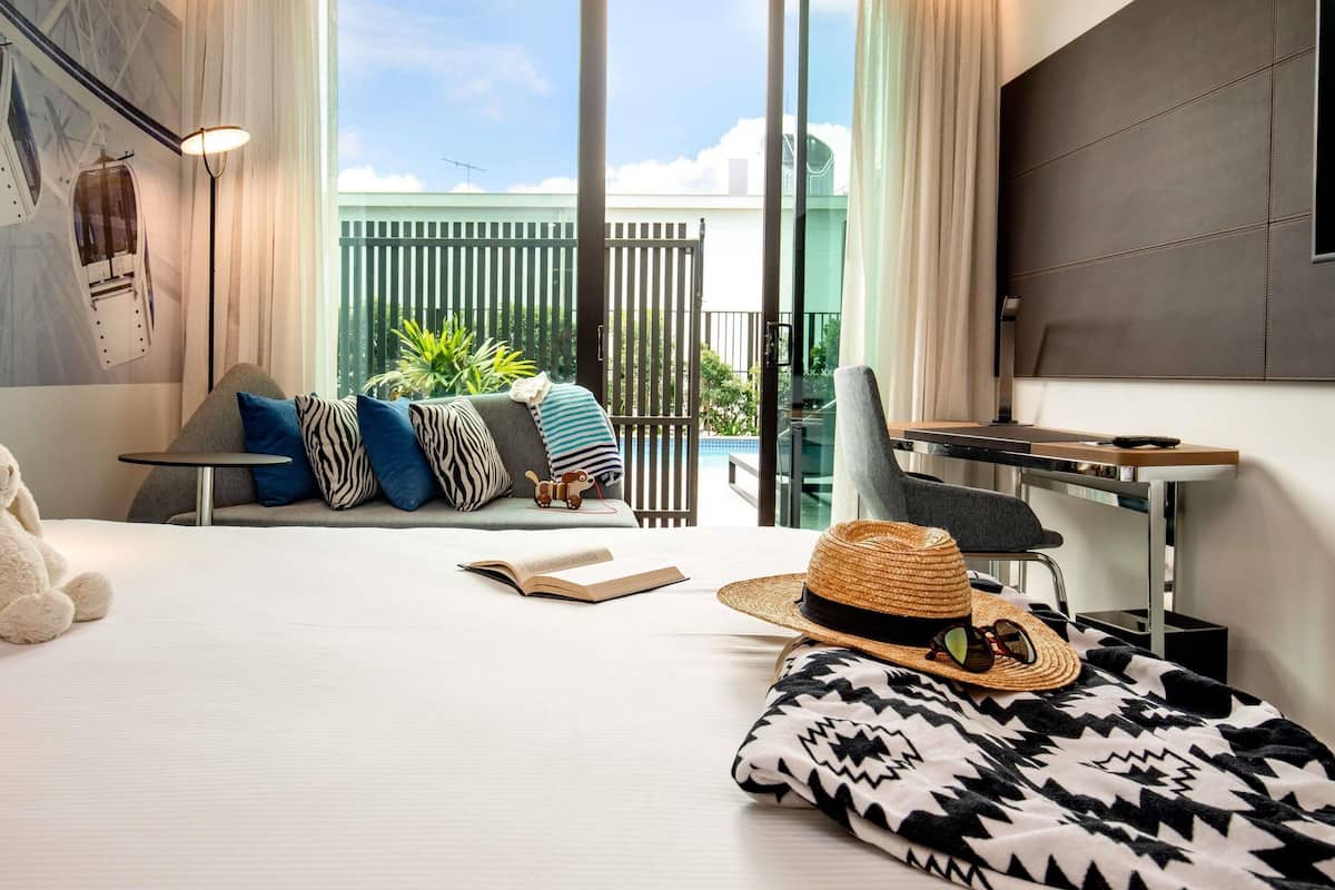 Una habitación de hotel moderna con una cama grande, un escritorio, una silla, un sofá con cojines estampados, un sombrero de paja y un libro.