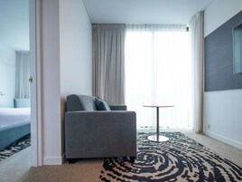 A hotel room with a grey sofa, a small round table, a patterned carpet, and a large window with curtains.