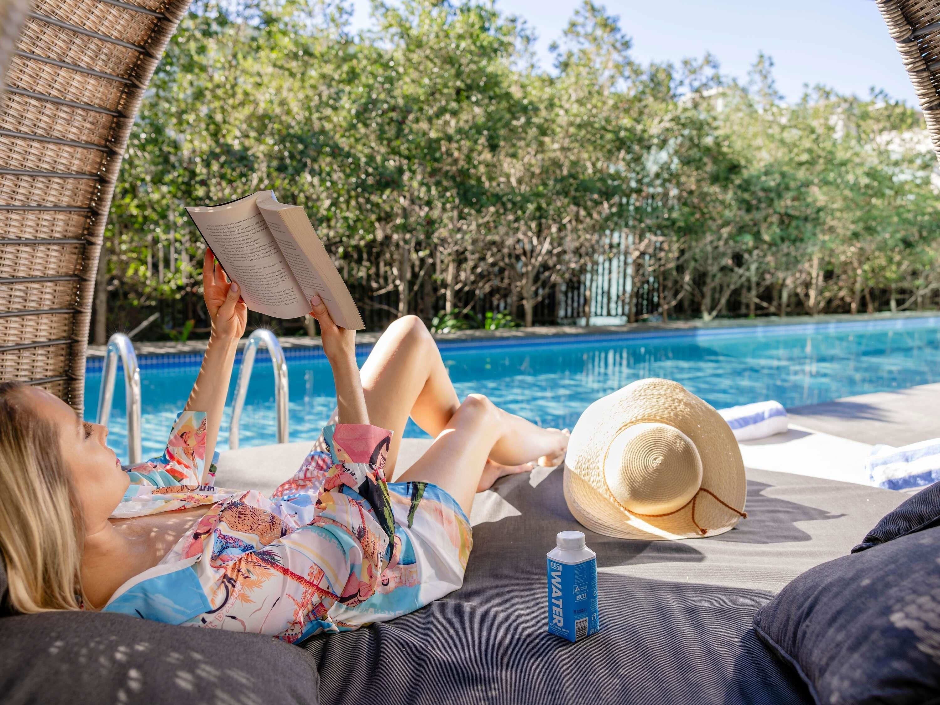 Una persona descansando junto a una tumbona en la piscina, leyendo un libro, con un sombrero de paja y una botella de agua cerca.