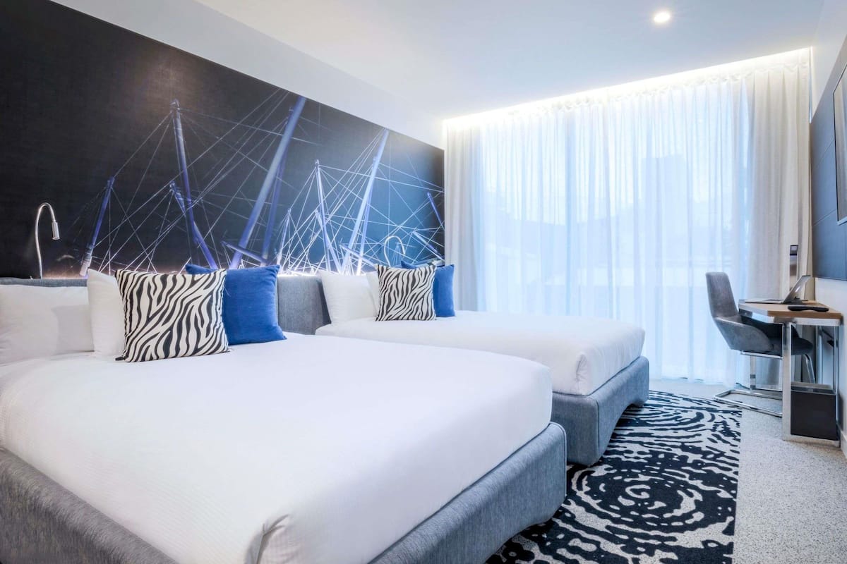 Una habitación de hotel moderna con una cama grande, dos mesitas de noche, un escritorio con silla y un ventanal con cortinas translúcidas.