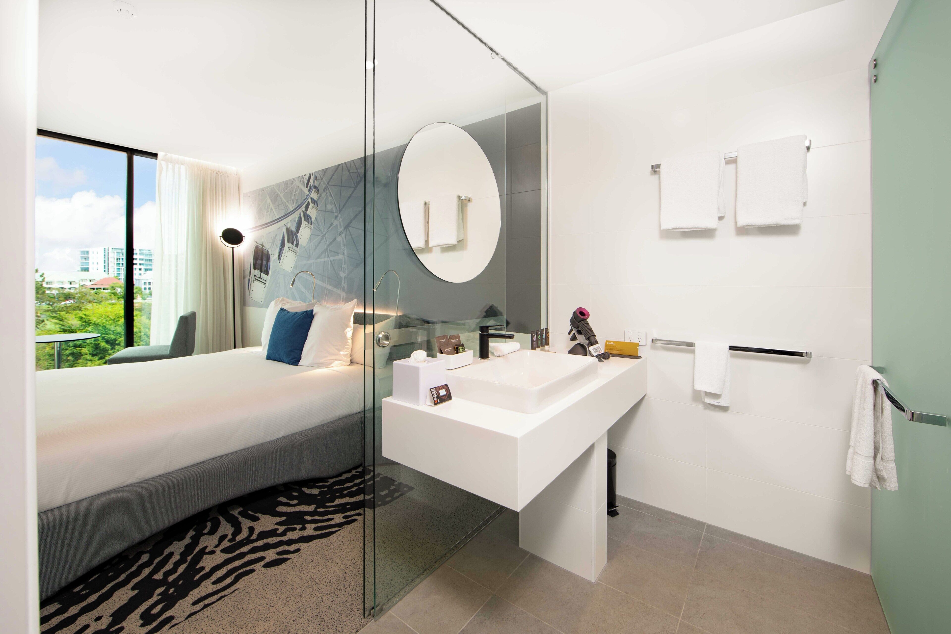 Un baño de hotel moderno con un amplio box de ducha de vidrio, un lavamanos central y una cama con vistas a la ciudad.
