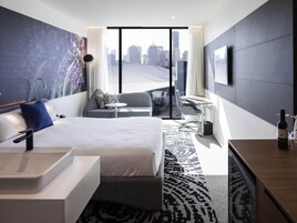 Una habitación de hotel moderna con una cama grande, un escritorio, una silla, una mesita y vistas a la silueta urbana.