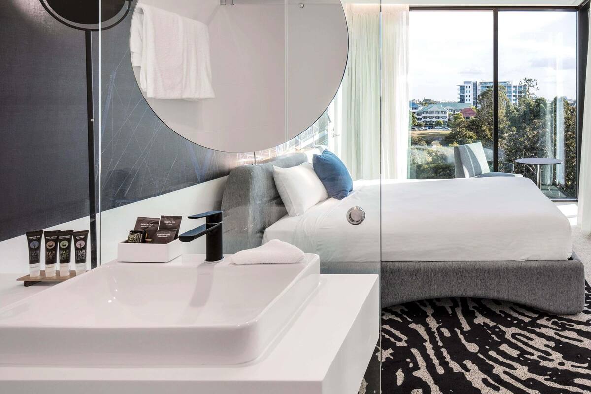Habitación de hotel moderna con una cama grande, un espejo redondo, un lavabo blanco y vistas a la ciudad a través de amplias ventanas.