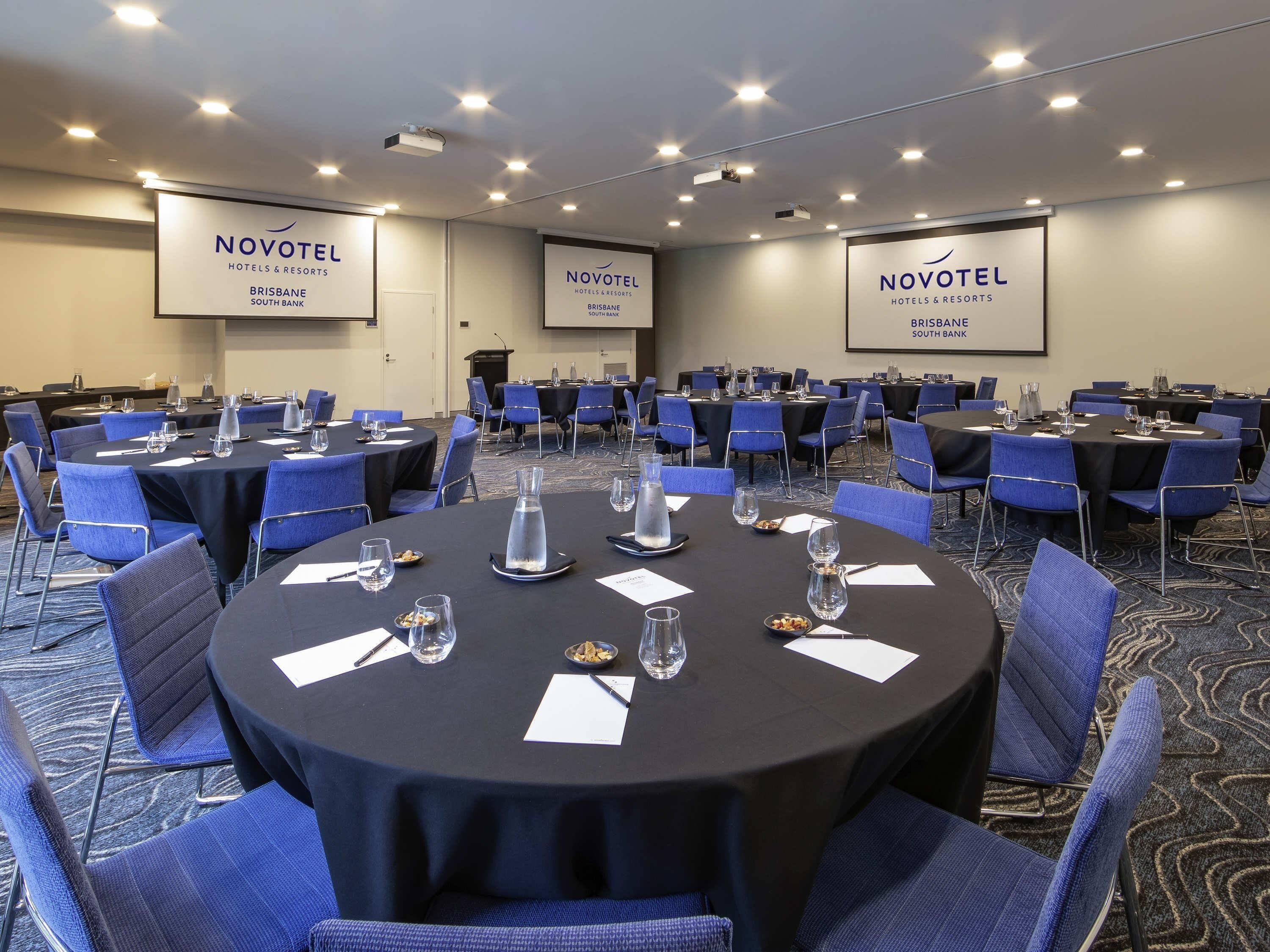 A conference room at Novotel Hotels & Resorts in Brisbane South Bank with round tables, blue chairs, and presentation screens.