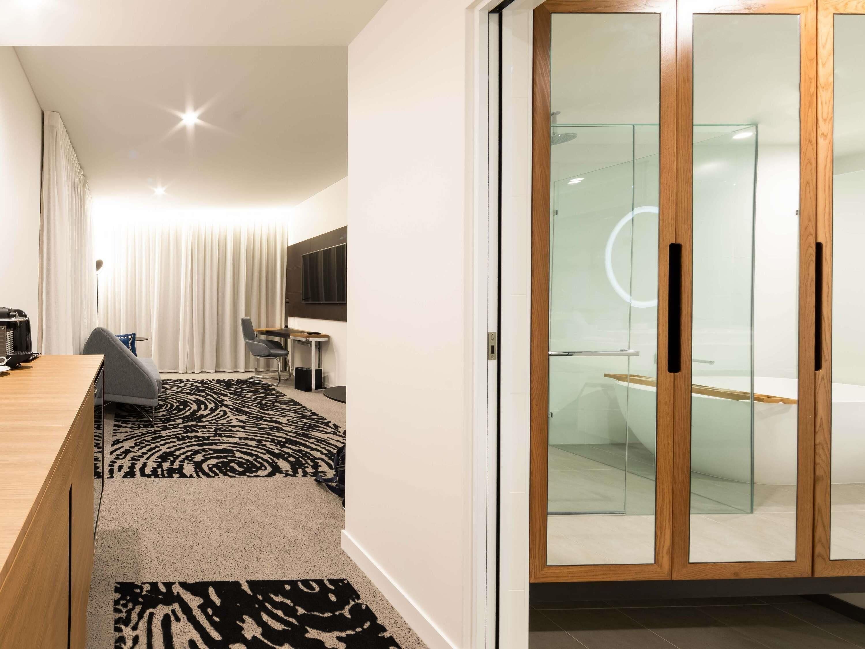 A modern hotel room with a wooden desk, a black and white patterned rug, a grey chair, a small table, a flat-screen TV, and a glass-enclosed shower area.