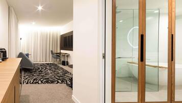 A modern hotel room with a wooden desk, a black and white patterned rug, a grey chair, a small table, a flat-screen TV, and a glass-enclosed shower area.