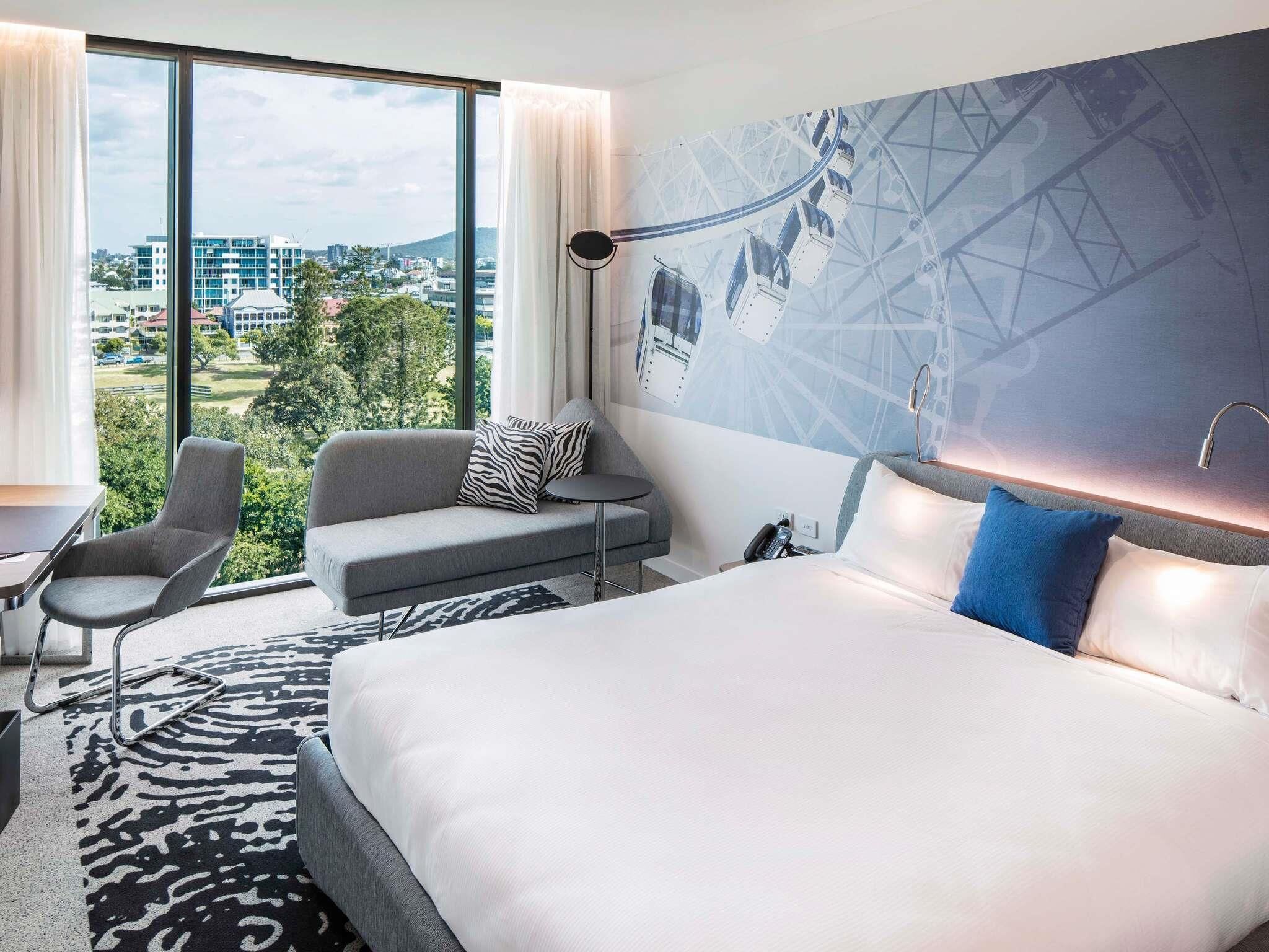 Moderni hotellihuone, jossa on suuri sänky, sohva, työpöytä ja suuret ikkunat, joista avautuu näkymä kaupungin ylle.