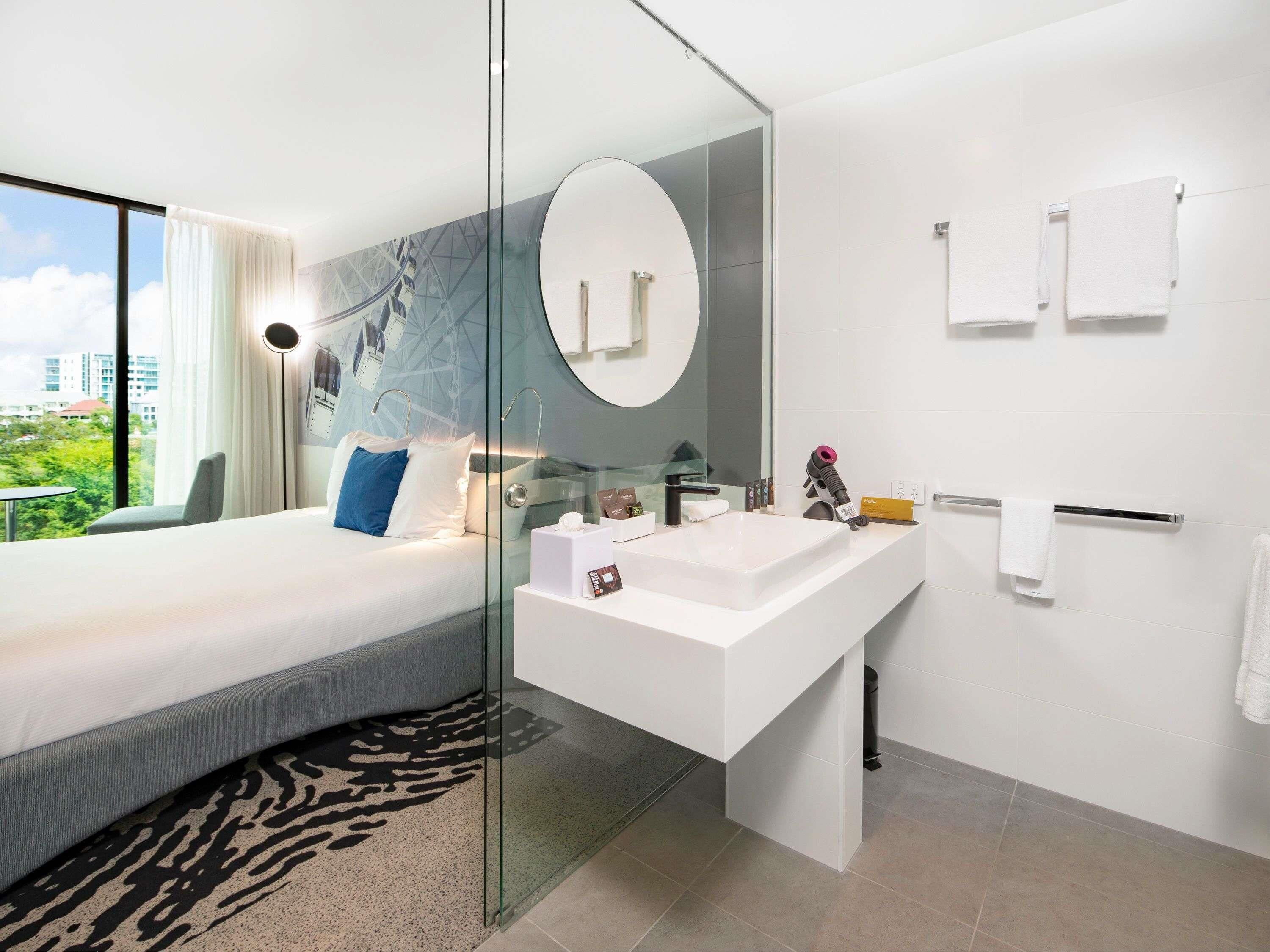 A modern hotel room with a bed, a large window, a glass-enclosed shower, a white sink, and a mirror.