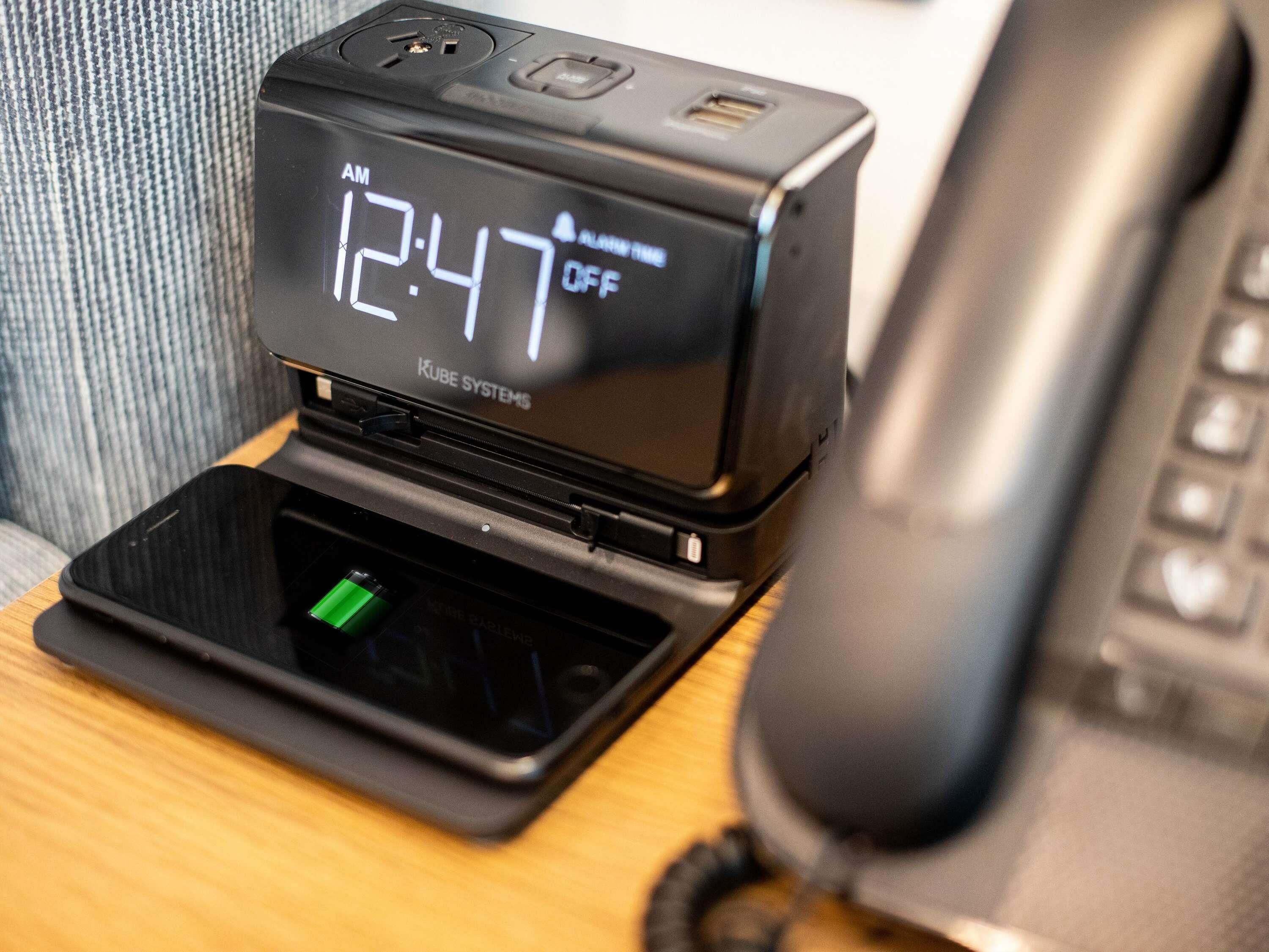 Un reloj digital muestra la 1:47 de la madrugada, con un botón de encendido y un puerto USB, junto a un teléfono sobre una superficie de madera.