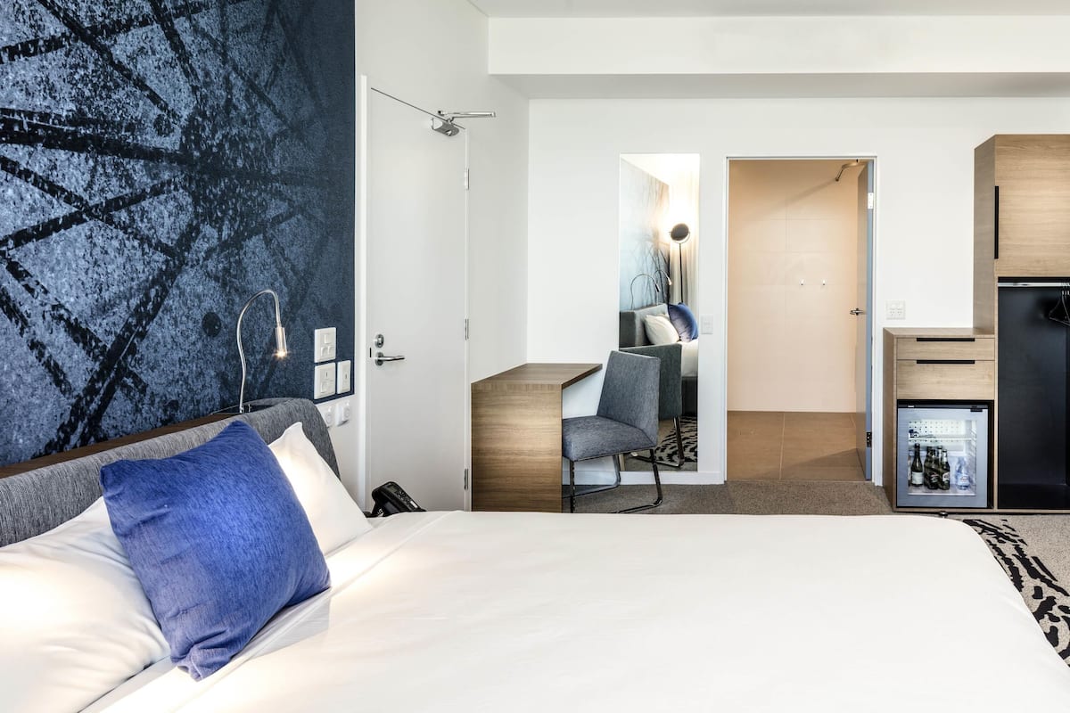 Una habitación de hotel moderna con una cama, un escritorio, una silla, un minibar y un baño visible a través de una puerta abierta.