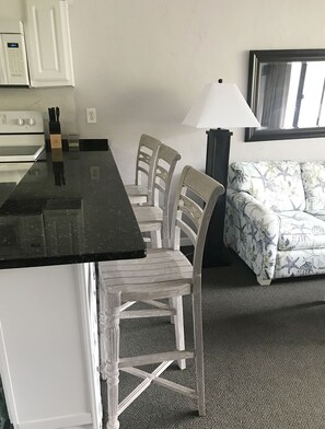 Une cuisine compacte avec un plan de travail noir, des placards blancs et un four encastré. On y trouve deux chaises blanches à ossature en bois et un canapé blanc à motifs floraux. Une lampe est placée sur le côté droit du canapé.