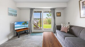 A living room with a TV on a wooden stand, a grey sofa with patterned cushions, and a sliding glass door leading to a garden.