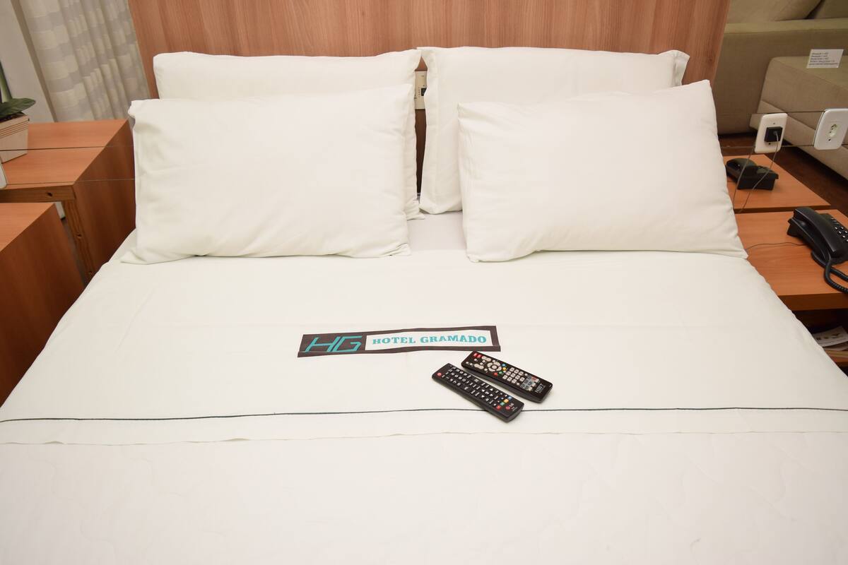 Un lit d’hôtel avec une literie blanche, deux télécommandes et une pancarte indiquant « HG HOTEL GRAMADO ».