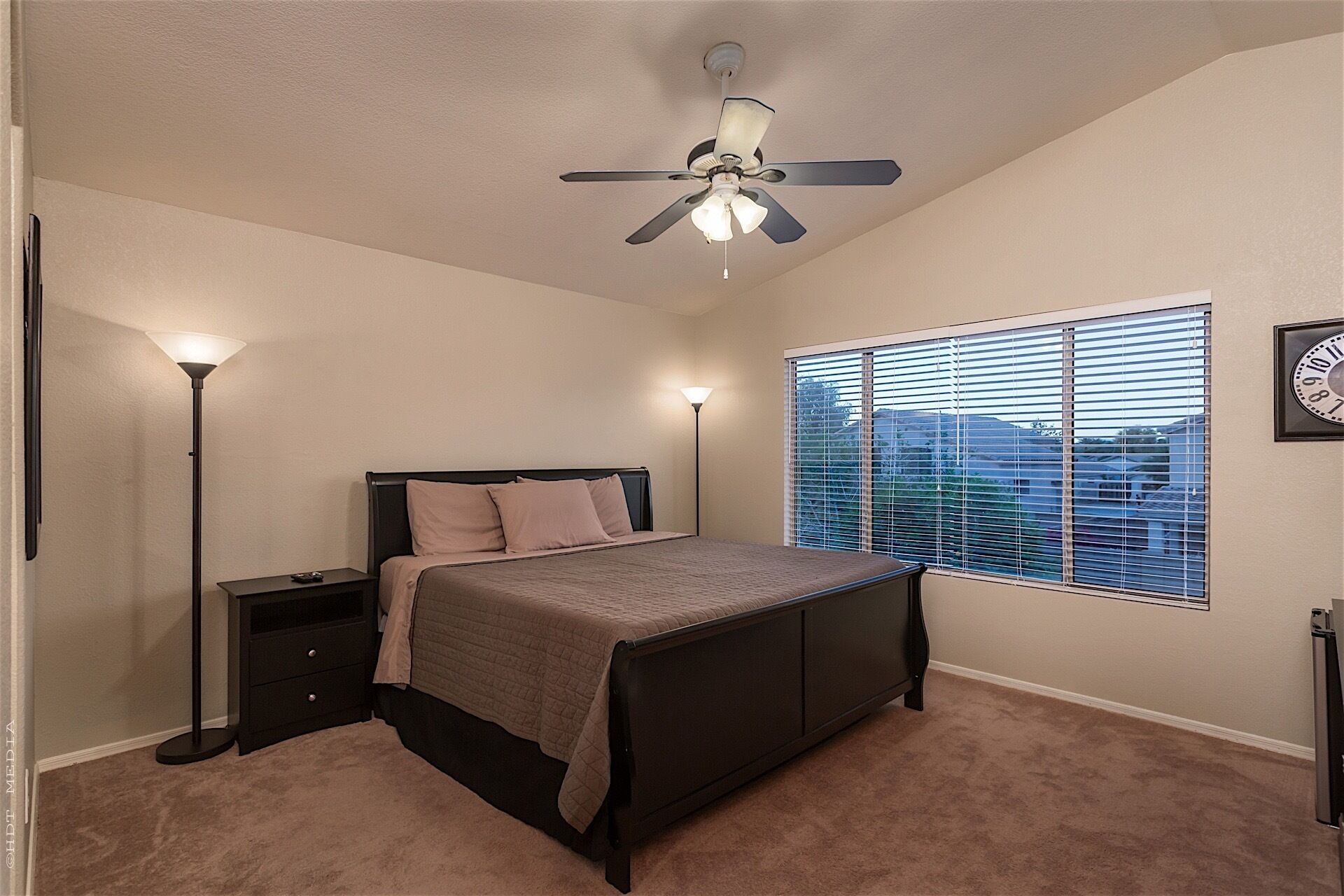 A bedroom with a bed, bedside lamps, a ceiling fan, a window with blinds, and a wall clock.