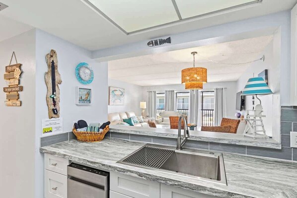 A modern kitchen with a built-in dishwasher, a stainless steel sink, and a large window overlooking a living area.