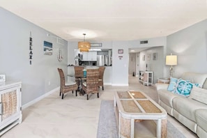 A modern living room with a sofa, a dining table with chairs, a kitchen in the background, and a hallway leading to another room.