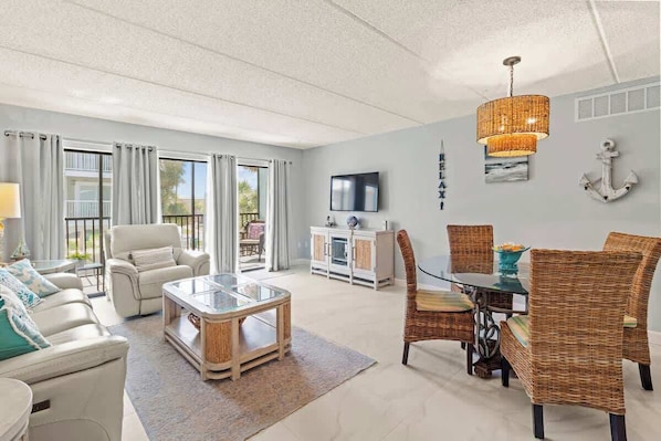 A spacious living room with a glass coffee table, wicker chairs, a TV, and a dining area with a table and chairs.