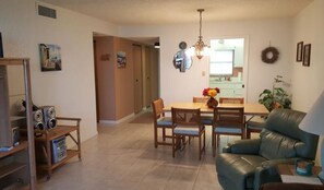Una sala de estar con un sofá de cuero, una mesa de comedor de madera y sillas. Se ve una cocina al fondo con un refrigerador y un lavabo.
