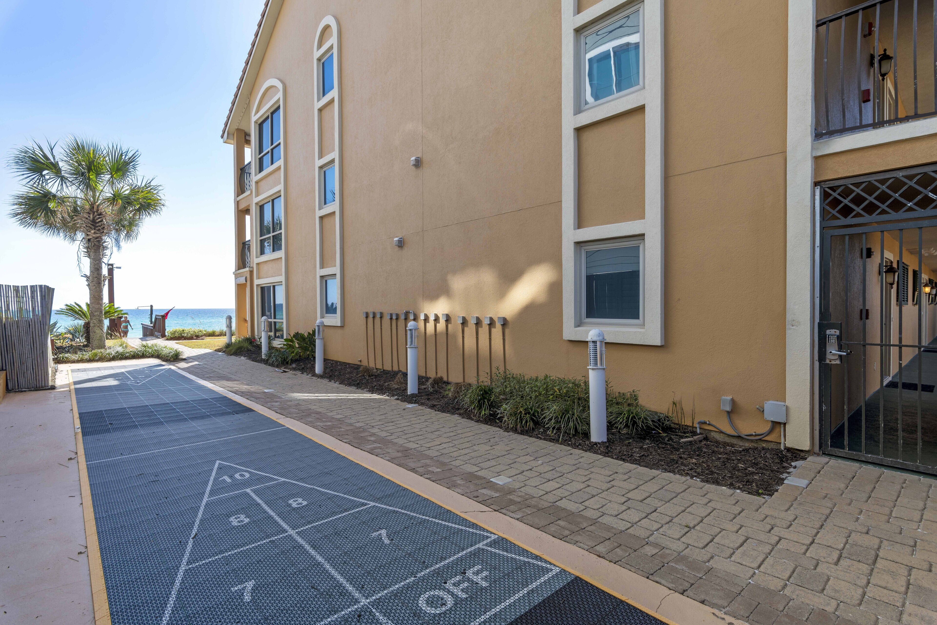 A building with a paved area marked for shuffleboard, a palm tree, and a view of the sea.