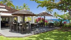 An outdoor dining area with thatched umbrellas, tables, and chairs, surrounded by palm trees and a view of the ocean. - Mauna Lani: Enjoy golf; snorkeling, helicopter tours Flexible cancellation terms (Kamuela)