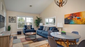 A modern living room with a blue sofa, a round coffee table, and a dining table with chairs. There are two windows with white frames, a ceiling light fixture, and a painting on the wall.