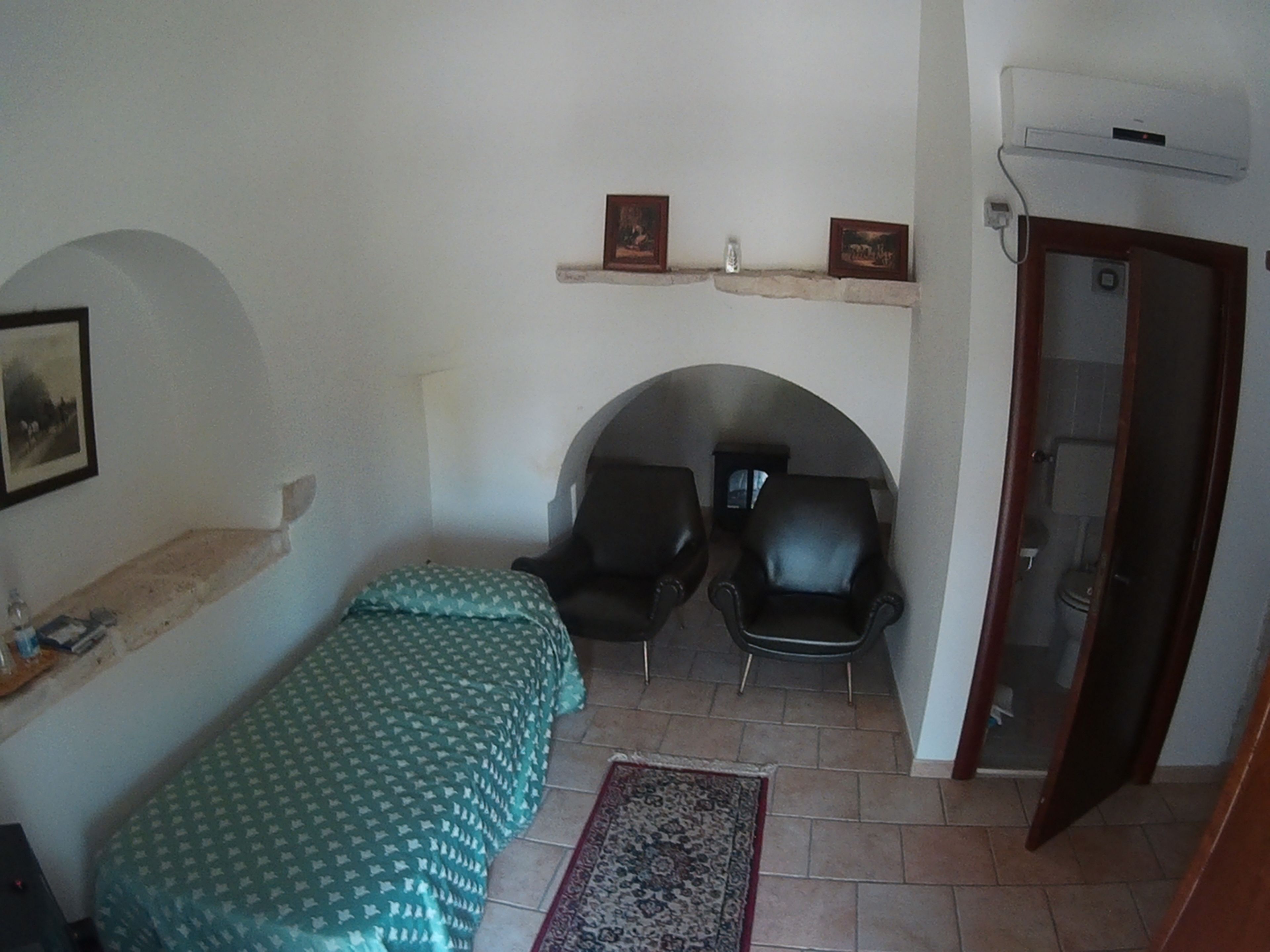 Une petite pièce, de taille modeste, comprenant un lit, deux chaises, une étagère avec des photos et une porte donnant sur une salle de bain.