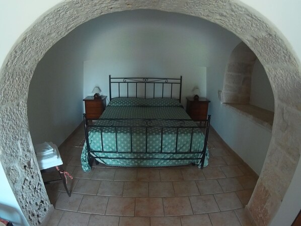 Quarto com cama sob um arco de pedra, duas mesas de cabeceira com luminárias e piso de azulejos.