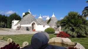 A traditional stone house with conical roofs, a stone wheel, and a well-maintained garden with red flowers and palm trees. - Tenuta Antico Leccio (Gioia del Colle)