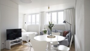 Una sala de estar moderna con un televisor de pantalla plana, una mesa de comedor redonda con sillas, un sofá y un gran ventanal con cortinas.