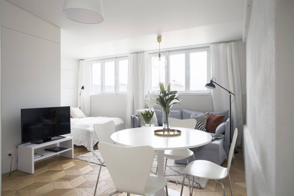 A modern living room with a flat-screen TV, a round dining table with chairs, a sofa, and a large window with curtains.