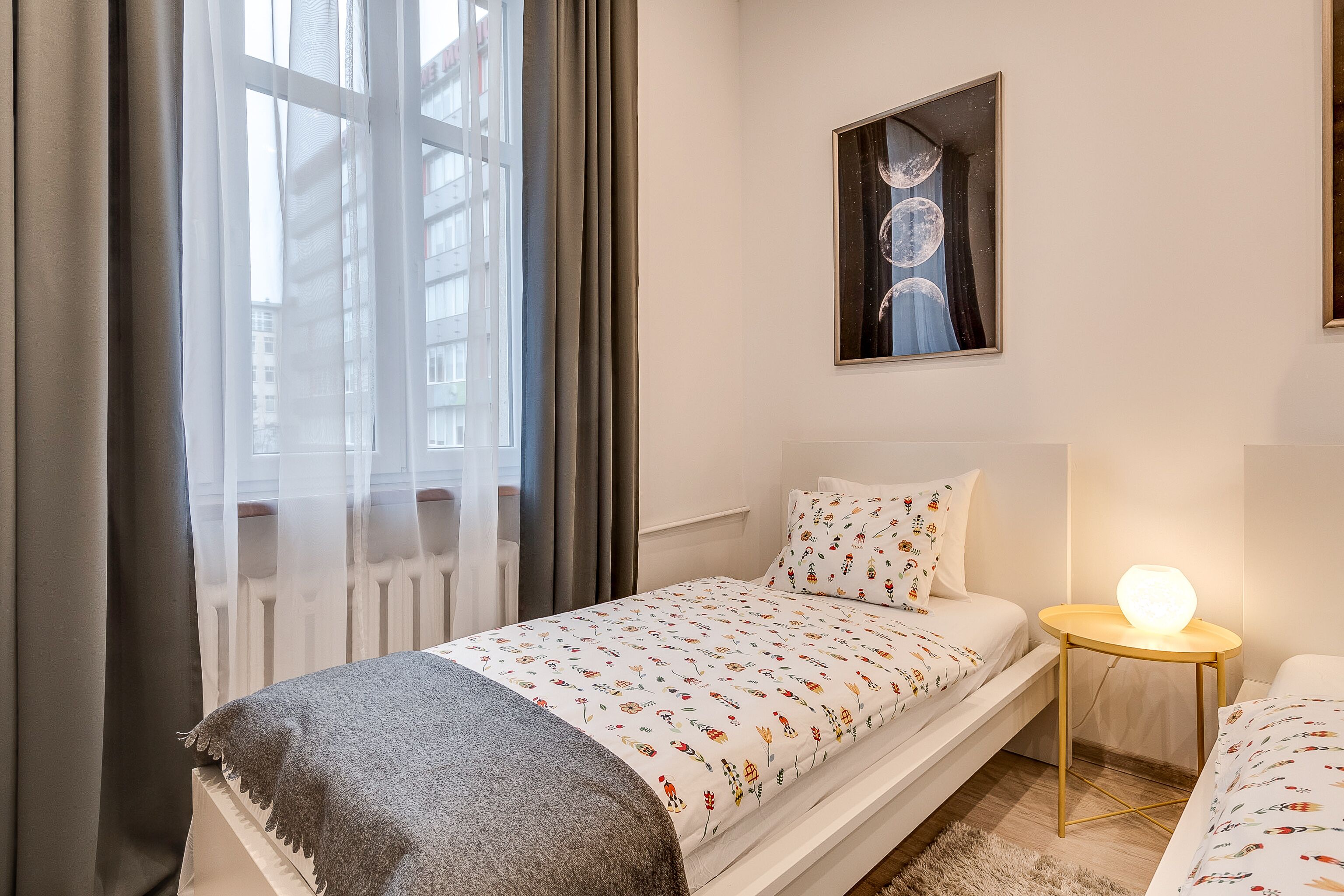 A modern bedroom with a bed, a bedside table with a lamp, a window with curtains, and a wall-mounted artwork.