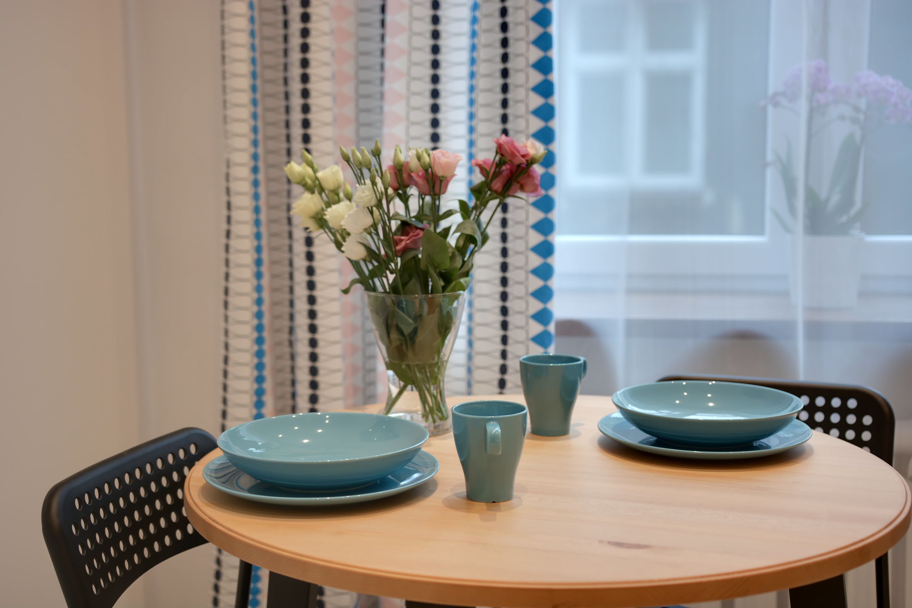 Una mesa de madera redonda con dos platos azules y una taza del mismo color, junto con un jarrón de flores.