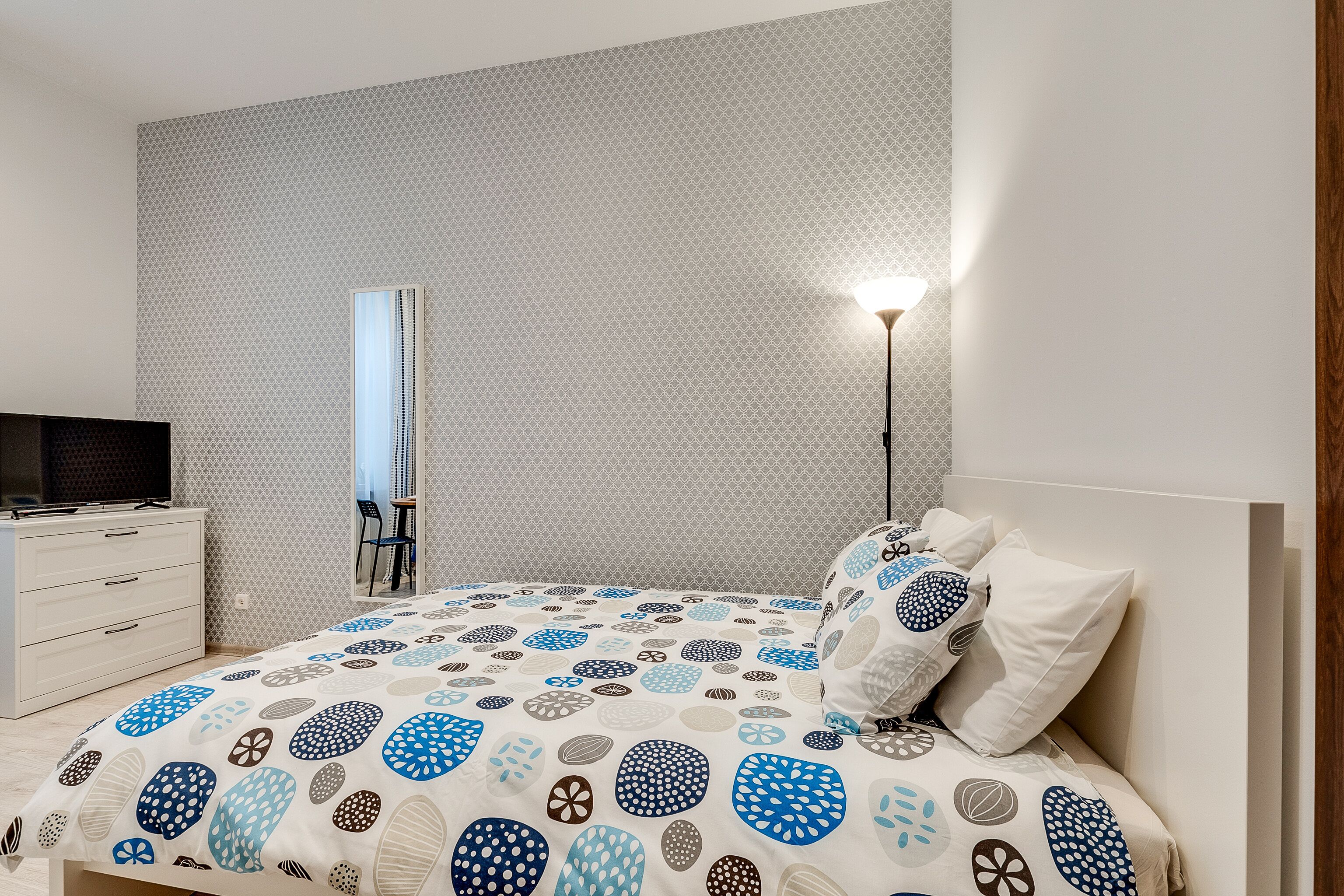 Un dormitorio con una colcha estampada, una lámpara de noche, un televisor en una cómoda y un espejo en la pared.