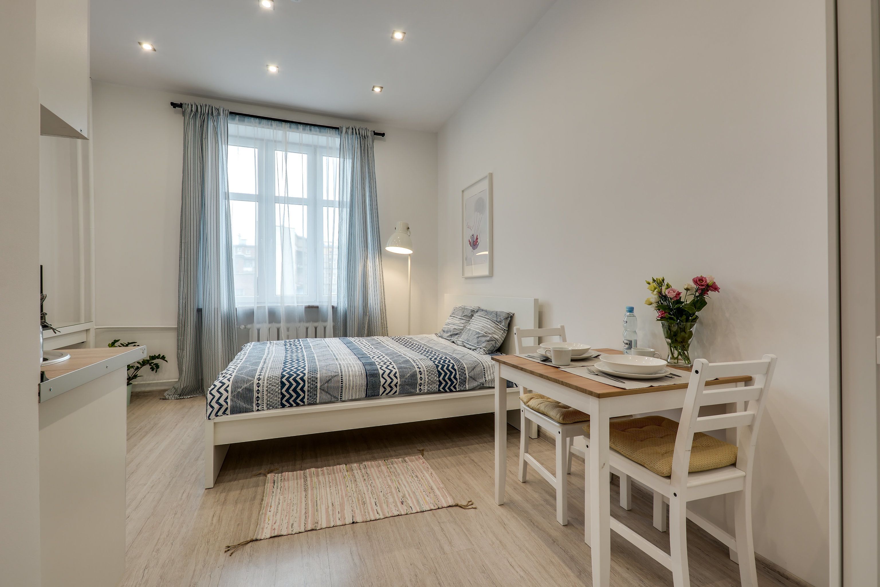 A bedroom with a bed, bedside table, lamp, and a small dining area with a table and chairs.