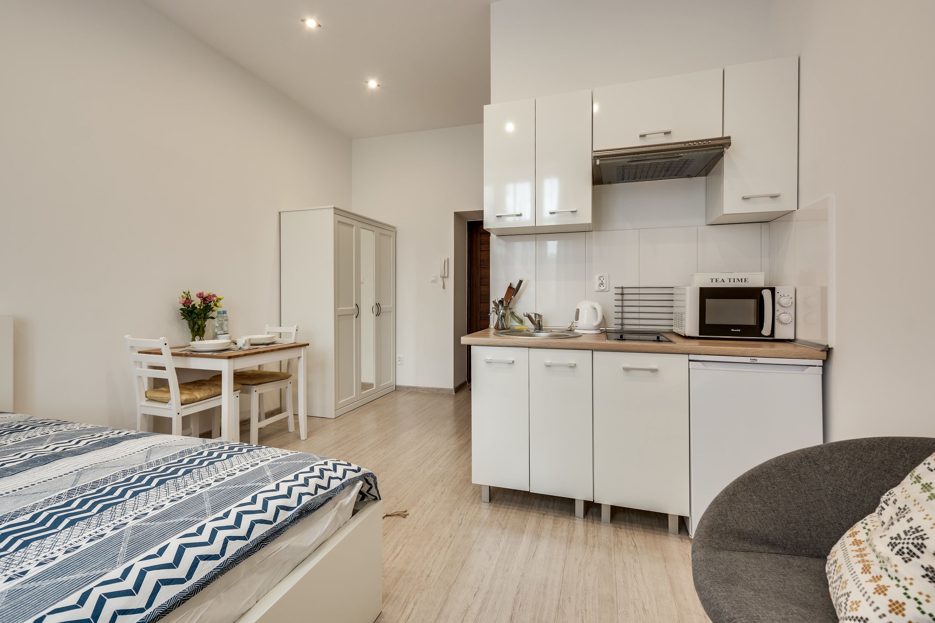 Un dormitorio compacto con cama, un escritorio pequeño y una kitchenette con microondas y hervidor de agua.