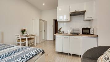 Un dormitorio compacto con cama, un escritorio pequeño y una kitchenette con microondas y hervidor de agua.