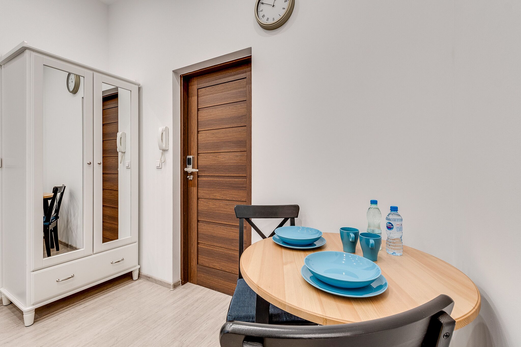 Ein Essbereich mit einem runden Holztisch, blauen Tellern und Stühlen. Es steht ein weißer Kleiderschrank mit Spiegel und eine Holztür mit einem Telefon dort.
